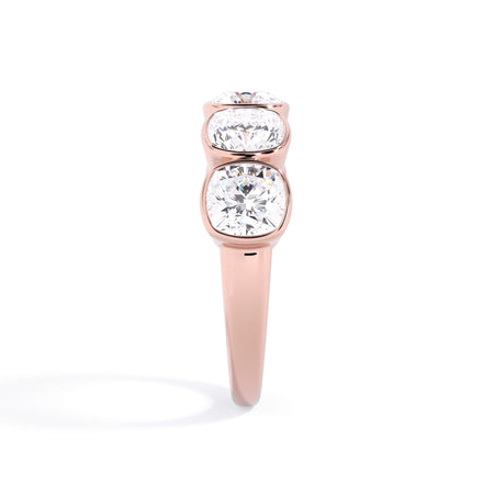 A picture of a five stone diamond wedding ring with cushion shape diamonds that are bezel set in rose gold on a white background with a side profile view.