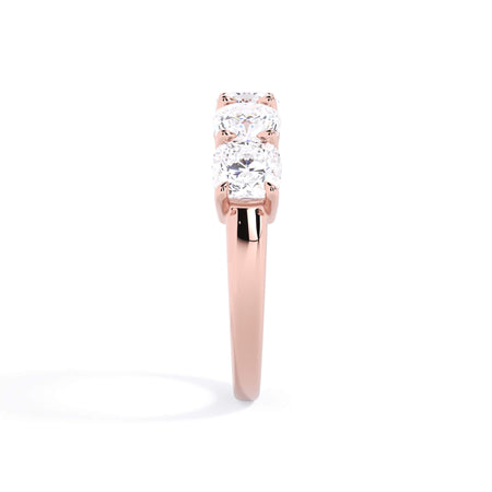 A picture of a five stone diamond wedding ring with cushion shape diamonds that are shared prong set in rose gold on a white background with a side profile view.