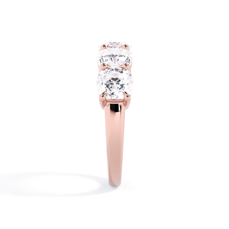 A picture of a five stone diamond wedding ring with cushion shape diamonds that are shared prong set in rose gold on a white background with a side profile view.