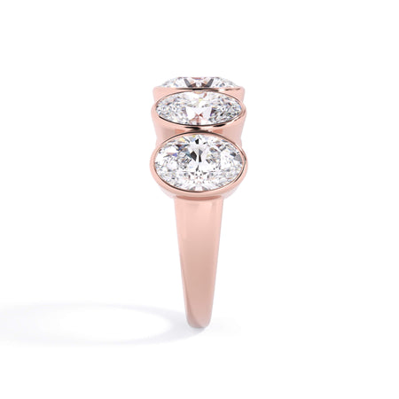 A picture of a five stone diamond wedding ring with oval shape diamonds that are bezel set in rose gold on a white background with a side profile view.