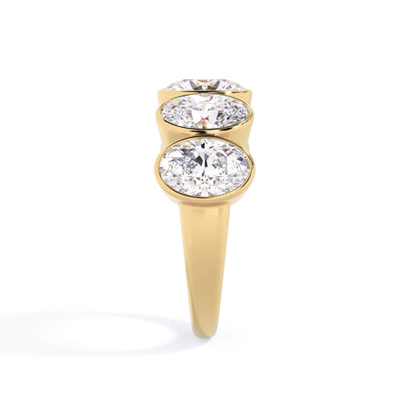 A picture of a five stone diamond wedding ring with oval shape diamonds that are bezel set in yellow gold on a white background with a side profile view.