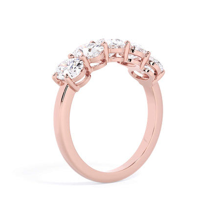 A picture of a five stone diamond wedding ring with oval shape diamonds that are shared prong set in rose gold on a white background with a slightly angled, through the finger view.