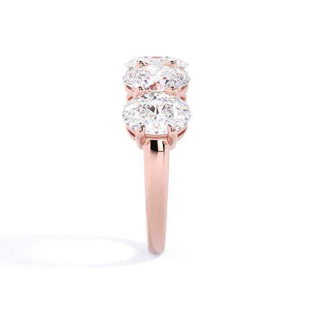 A picture of a five stone diamond wedding ring with oval shape diamonds that are shared prong set in rose gold on a white background with a side profile view.