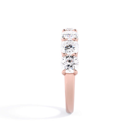 A picture of a seven stone diamond wedding ring with cushion cut diamonds that are shared prong set in rose gold on a white background with a side profile view.