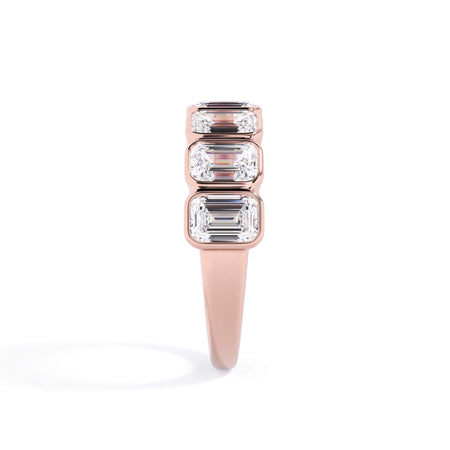 A picture of a seven stone diamond wedding ring with emerald cut diamonds that are bezel set in rose gold on a white background with a side profile view.