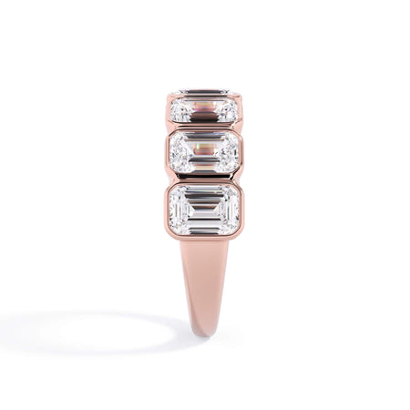 A picture of a seven stone diamond wedding ring with emerald cut diamonds that are bezel set in rose gold on a white background with a side profile view.