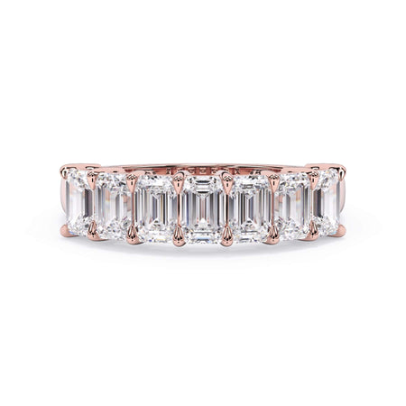 A picture of a seven stone diamond wedding ring with emerald cut diamonds that are shared prong set in rose gold on a white background with the ring facing forward, lying down.