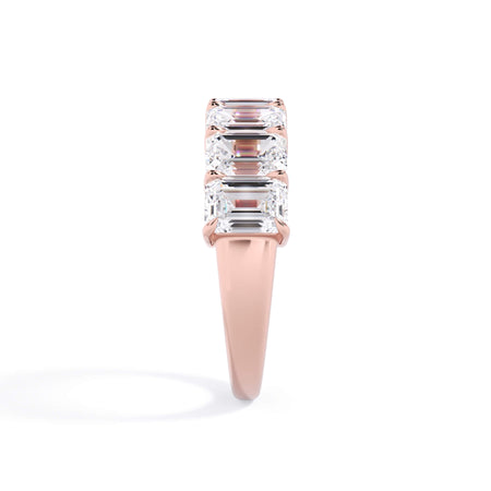A picture of a seven stone diamond wedding ring with emerald cut diamonds that are shared prong set in rose gold on a white background with a side profile view.