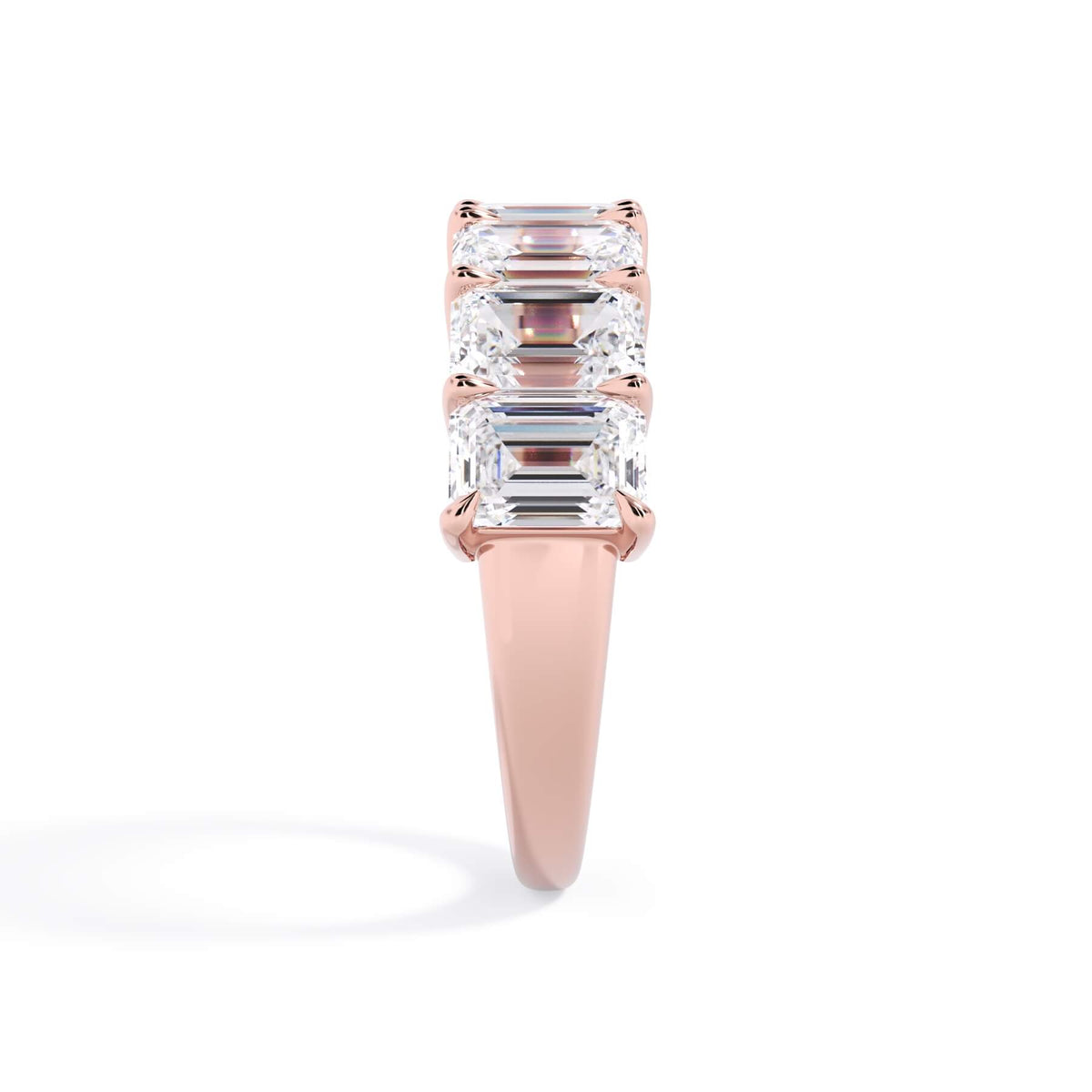A picture of a seven stone diamond wedding ring with emerald cut diamonds that are shared prong set in rose gold on a white background with a side profile view.