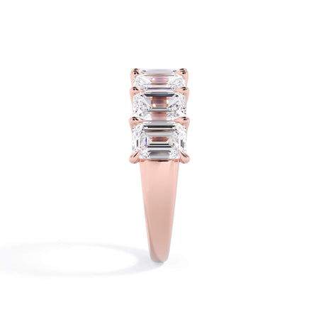 A picture of a seven stone diamond wedding ring with emerald cut diamonds that are U prong set in rose gold on a white background with a side profile view.