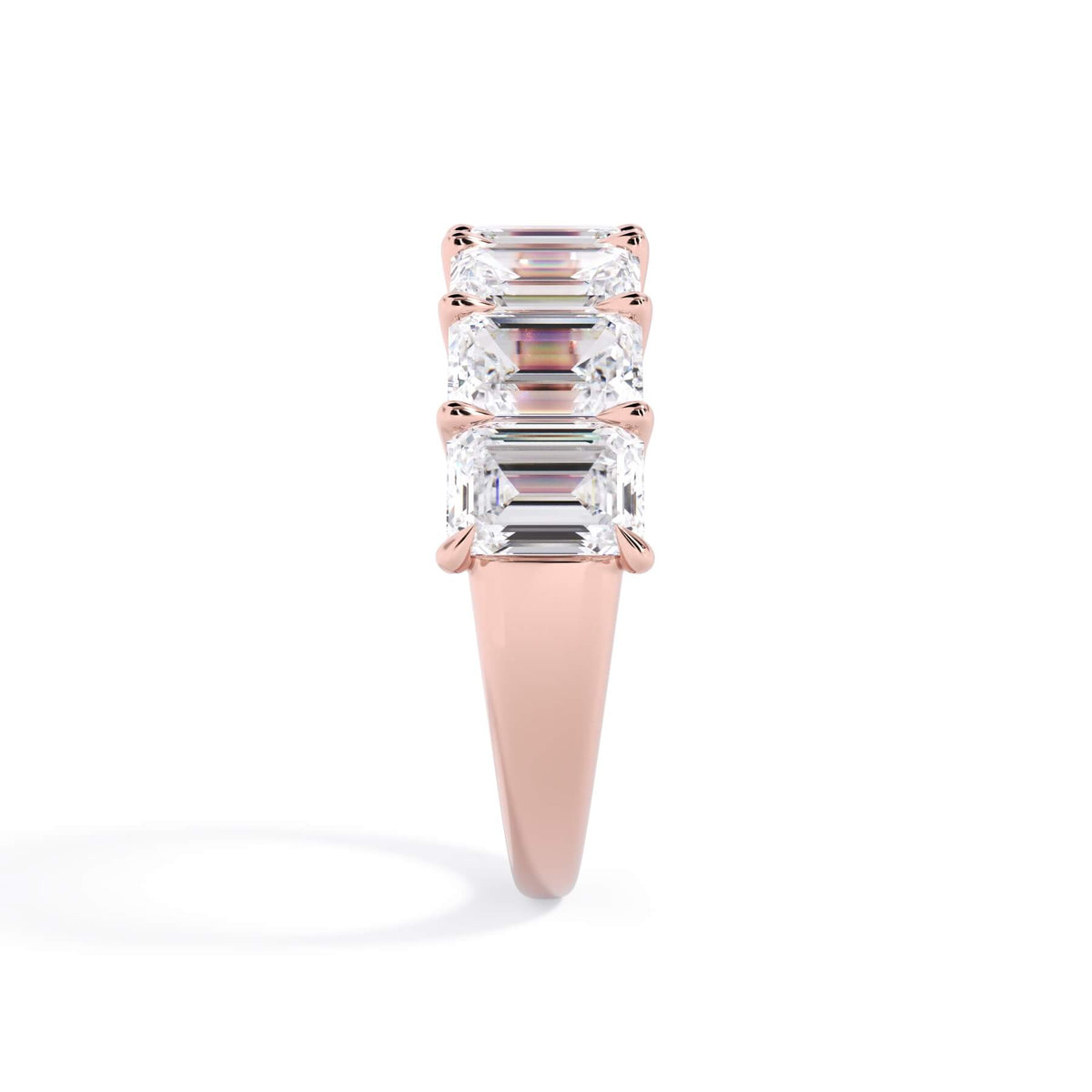 A picture of a seven stone diamond wedding ring with emerald cut diamonds that are U prong set in rose gold on a white background with a side profile view.