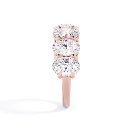 A picture of a seven stone diamond wedding ring with oval shaped diamonds that are shared prong set in rose gold on a white background with a side profile view.
