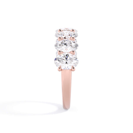 A picture of a seven stone diamond wedding ring with oval shaped diamonds that are U prong set in rose gold on a white background with a side profile view.