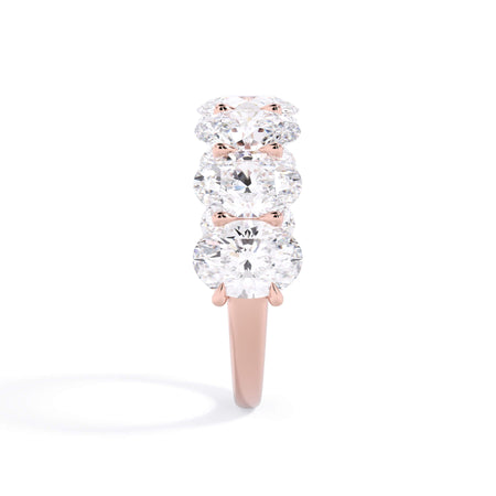 A picture of a seven stone diamond wedding ring with oval shaped diamonds that are U prong set in rose gold on a white background with a side profile view.