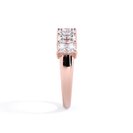 A picture of a seven stone diamond wedding ring with radiant cut diamonds that are shared prong set in rose gold on a white background with a side profile view.