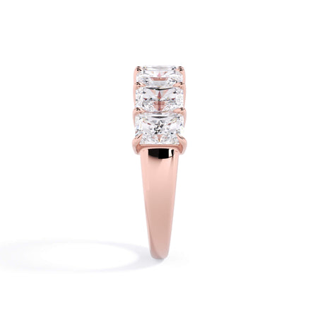 A picture of a seven stone diamond wedding ring with radiant cut diamonds that are shared prong set in rose gold on a white background with a side profile view.