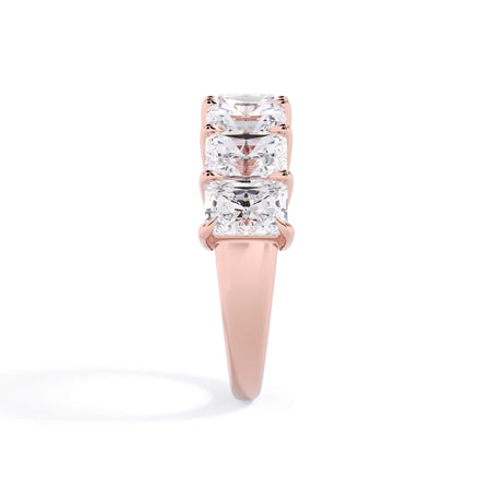 A picture of a seven stone diamond wedding ring with radiant cut diamonds that are shared prong set in rose gold on a white background with a side profile view.