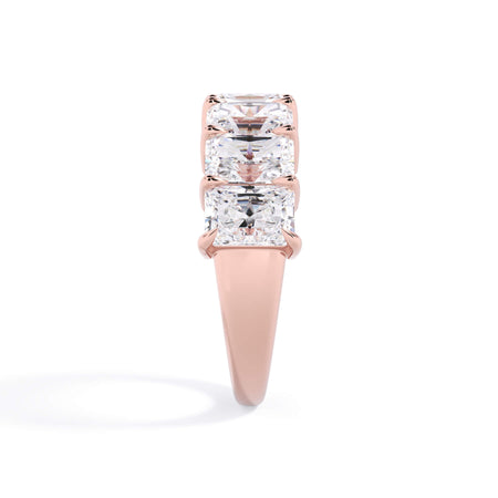 A picture of a seven stone diamond wedding ring with radiant cut diamonds that are shared prong set in rose gold on a white background with a side profile view.