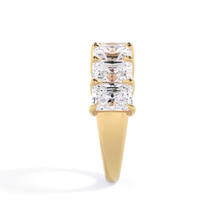 A picture of a seven stone diamond wedding ring with radiant cut diamonds that are shared prong set in yellow gold on a white background with a side profile view.
