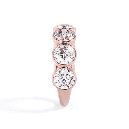A picture of a seven stone diamond wedding ring with round diamonds that are bezel set in rose gold on a white background with a side profile view.