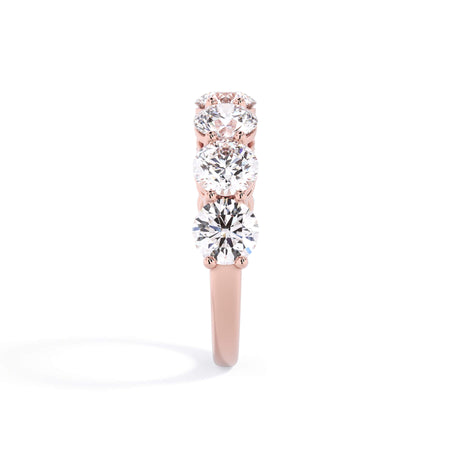 A picture of a seven stone diamond wedding ring with round diamonds that are shared prong set in rose gold on a white background with a side profile view.