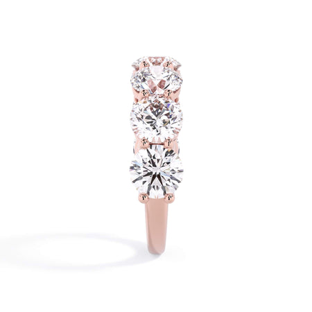 A picture of a seven stone diamond wedding ring with round diamonds that are shared prong set in rose gold on a white background with a side profile view.