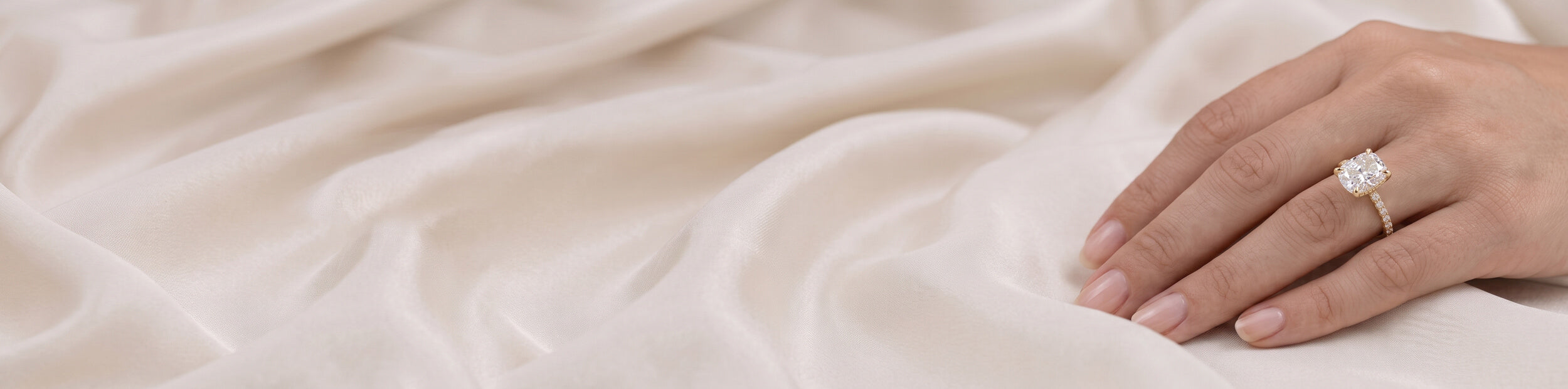 A close-up of a hand wearing a diamond engagement ring, resting on draped cream-colored satin fabric.