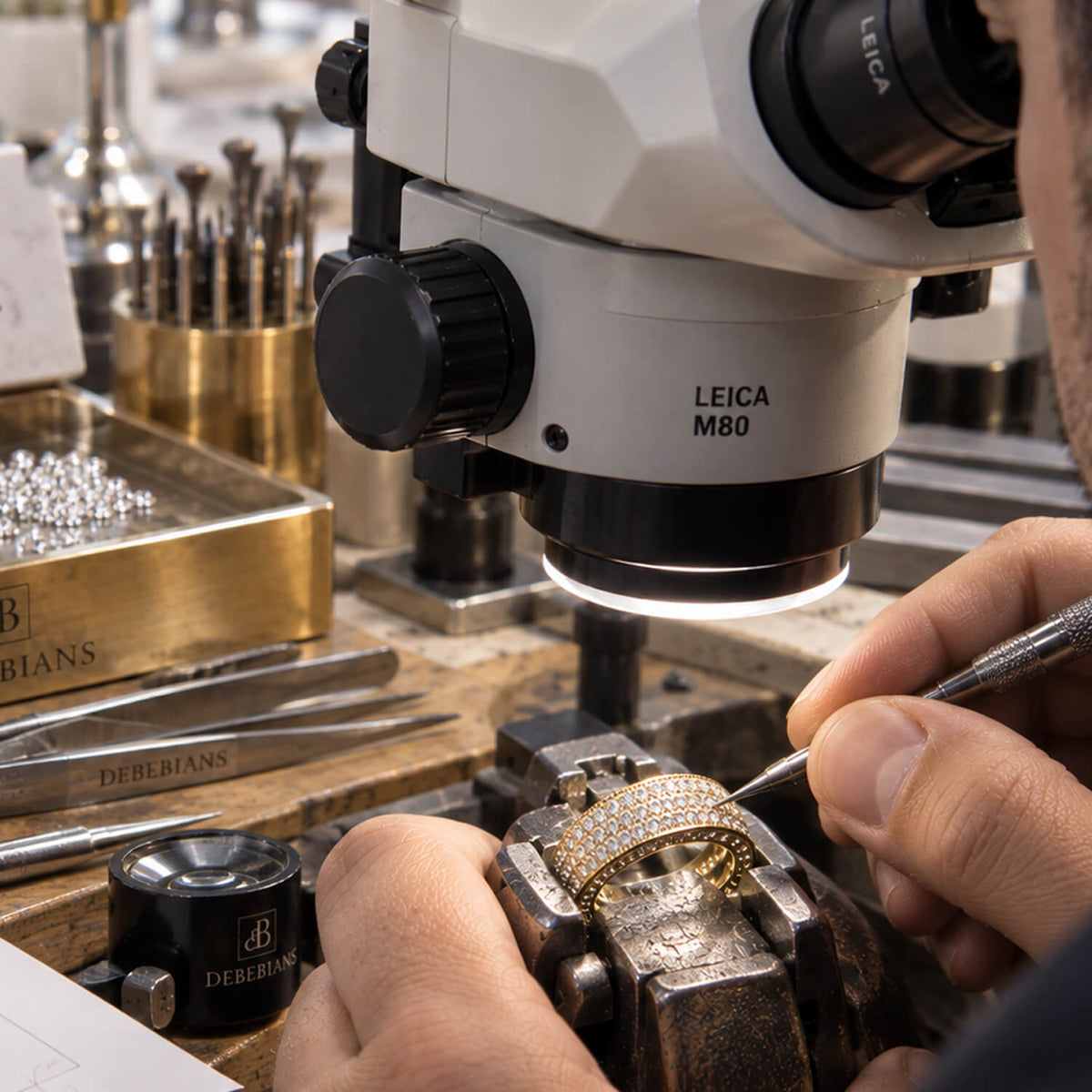 A close-up of a jeweler working under a Leica M80 microscope, carefully setting a gold ring studded with small clear stones. Hands, tools, and jewelry supplies are visible on a workbench in a detailed workshop setting.