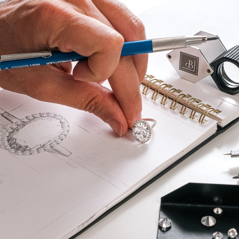 Jeweler holding engagement ring and pencil on top of a sketch pad with diamonds in the foreground.