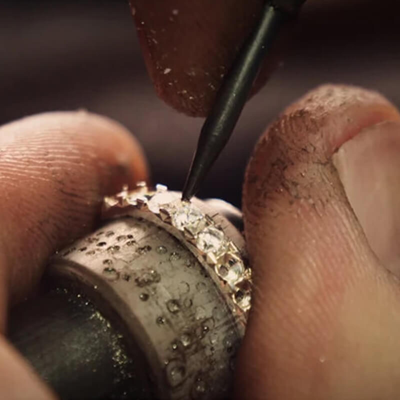 A close-up of the hand of a jeweler setting a diamond into a gold eternity ring.